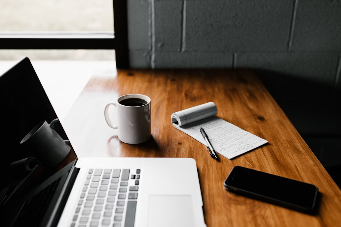 a phone,cup,laptop,note and pen on a table